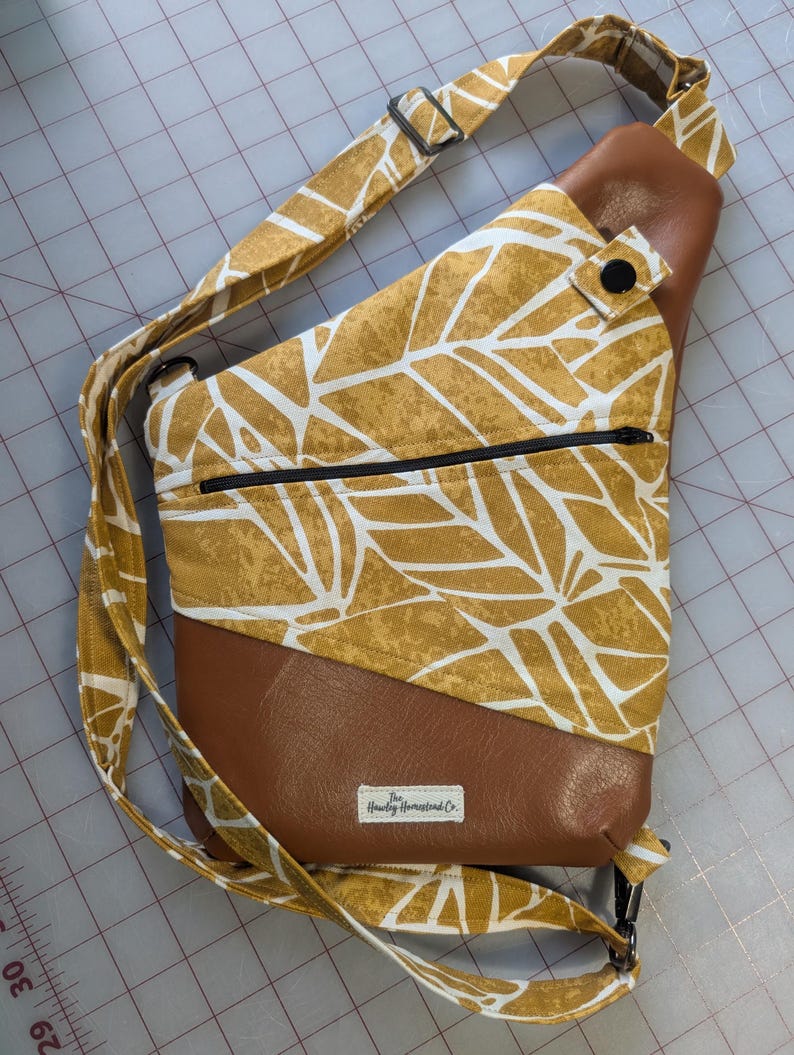 Op de afbeelding: Een mosterdgele sling bag met een wit bladerenpatroon en bruine, lederachtige accenten. De tas heeft een zwarte ritssluiting, een verstelbare riem en een label met "Hawley Homestead Co."