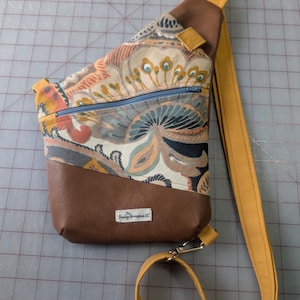 Op de afbeelding: Een sling bag met een bruine kunstleren basis en een bovenkant van stof met een patroon. De stof heeft een mix van kleuren, waaronder oranje, blauw en geel. De tas heeft een mosterdgele riem en een blauwe rits.