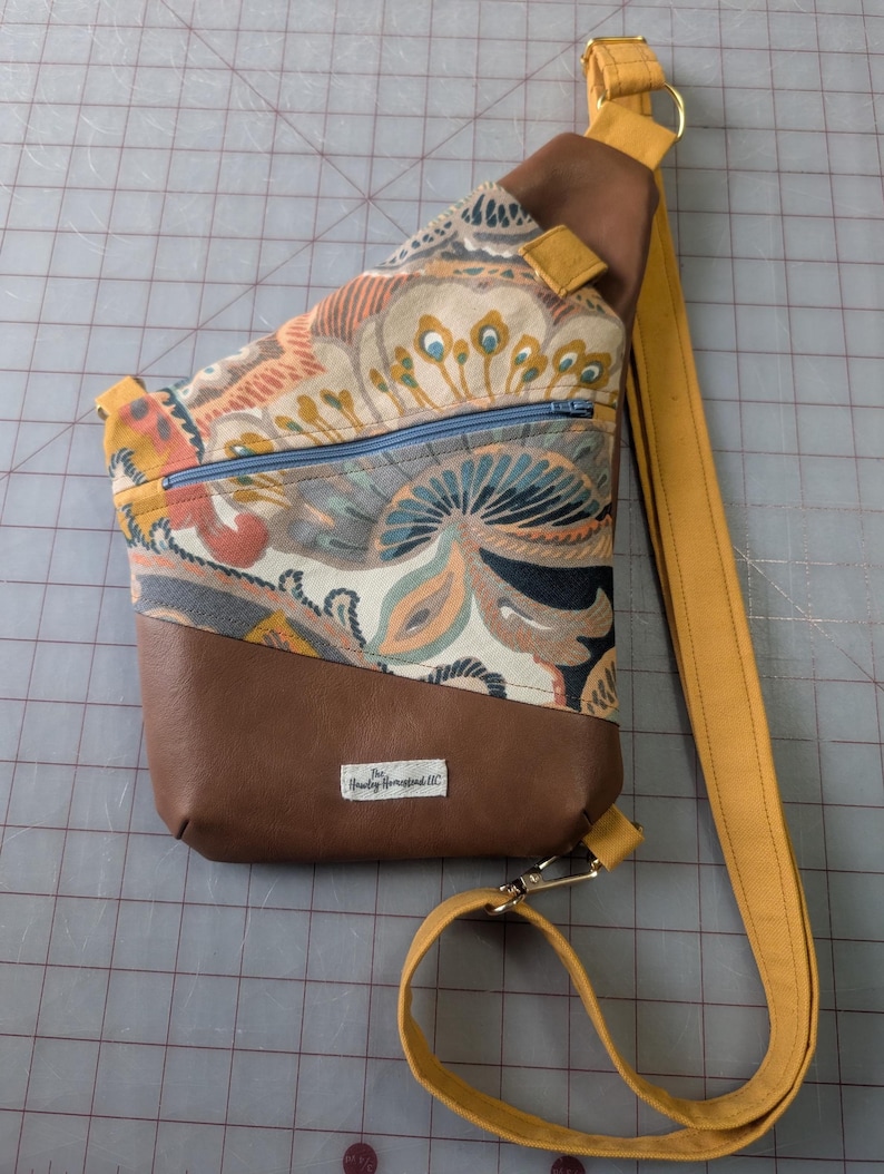Op de afbeelding: Een sling bag met een bruine kunstleren basis en een bovenkant van stof met een patroon. De stof heeft een mix van kleuren, waaronder oranje, blauw en geel. De tas heeft een mosterdgele riem en een blauwe rits.