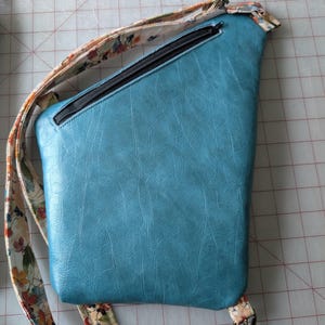 Op de afbeelding: Een turquoise crossbodytas met een zwarte rits en een bloemenpatroonriem. De tas heeft een getextureerd oppervlak en een rechthoekige vorm met een diagonale bovenkant. De riem is cr&egrave;mekleurig met een kleurrijk bloemmotief.