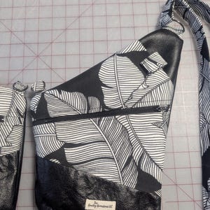 Op de afbeelding: Zwarte en witte sling bag met een tropisch bladerpatroon. De tas heeft een zwarte leren basis en een vak met ritssluiting. De riem heeft ook het bladerpatroon. De tekst "The Howdy Homestead LLC" is zichtbaar.