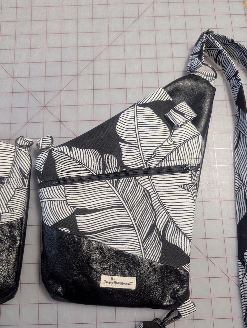 Op de afbeelding: Zwarte en witte sling bag met een tropisch bladerpatroon. De tas heeft een zwarte leren basis en een vak met ritssluiting. De riem heeft ook het bladerpatroon. De tekst "The Howdy Homestead LLC" is zichtbaar.