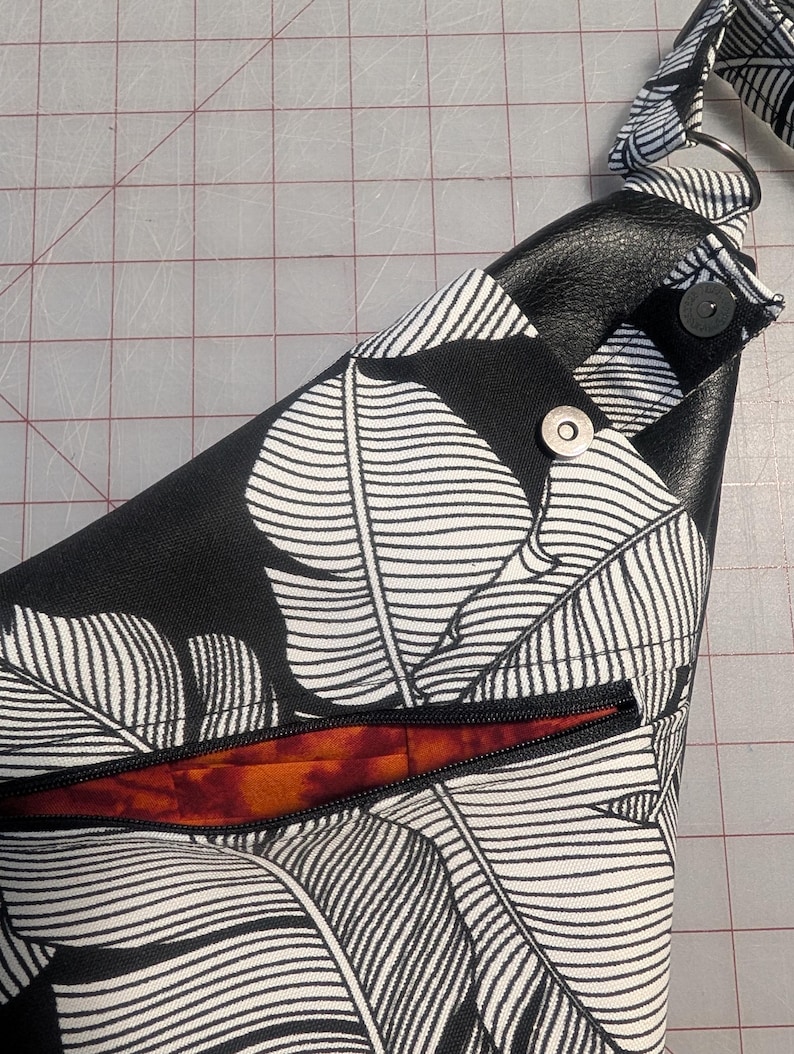 Op de afbeelding: Schoudertas met een zwart-wit bladpatroon. De tas heeft een zwarte rits, een zwart lederen detail en een magnetische sluiting. De binnenvoering is oranje en rood.