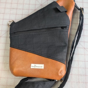 Op de afbeelding: Een zwarte en bruine sling bag met een gewatteerde zwarte stoffen bovenkant en een bruine imitatie leren onderkant. De tas heeft een zwarte verstelbare riem, een vak met ritssluiting en een klein label met "The Hawley Homestead LLC".