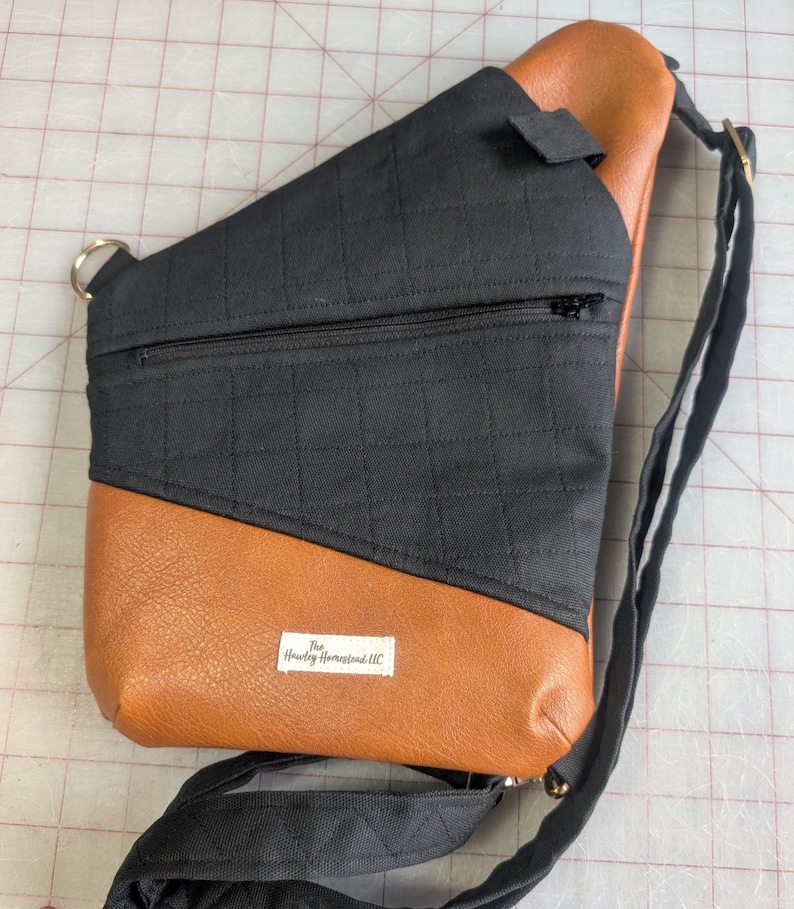 Op de afbeelding: Een zwarte en bruine sling bag met een gewatteerde zwarte stoffen bovenkant en een bruine imitatie leren onderkant. De tas heeft een zwarte verstelbare riem, een vak met ritssluiting en een klein label met "The Hawley Homestead LLC".