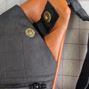 Op de afbeelding: Close-up van een zwarte en bruine sling bag. De tas heeft een zwart stoffen gedeelte met een ritssluiting en een drukknoopsluiting. De hoofdbody is van bruin leerachtig materiaal. Een zwarte riem is zichtbaar.
