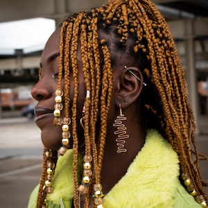 Pode incluir: Uma mulher com longas tranças marrons com contas douradas e um brinco dourado. Ela está vestindo uma jaqueta verde com gola peluda.