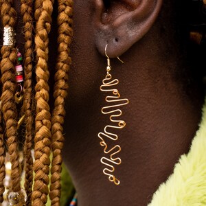Pode incluir: Um close-up de uma orelha de uma pessoa com um brinco longo de fio dourado. O brinco tem a forma de uma série de linhas onduladas. A pessoa tem cabelo trançado castanho.