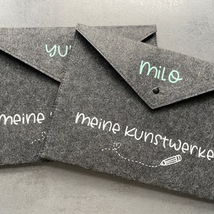 Könnte beinhalten: Drei graue Filzordner mit weißem Text. Der obere Ordner sagt "Milo" und "meine Kunstwerke" mit einer Zeichnung eines Bleistifts. Der mittlere Ordner sagt "Yu" und der untere Ordner sagt "meine".