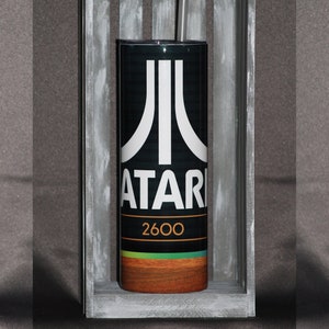 May include: A black and white tumbler with a retro Atari 2600 logo and a rainbow stripe design. The tumbler is in a wooden crate.