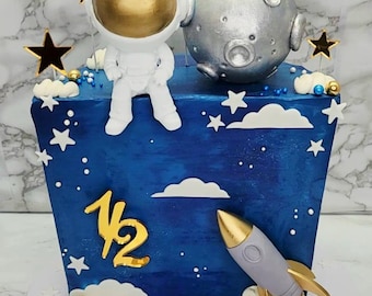 May include: A blue cake decorated with an astronaut, a silver moon, and a gold rocket. The cake is adorned with gold stars, clouds, and the number "1/2". The name "ATLAS" is written on the base in gold letters. A festive cake for a space-themed celebration.