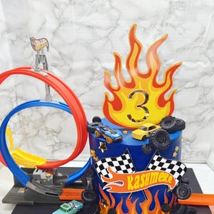 May include: A blue birthday cake decorated with toy cars, checkered flags, and a flame-shaped number three. The cake has a name banner and is surrounded by toy car tires and a race track.