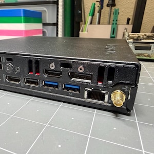 Puede incluir: Un dispositivo informático negro con múltiples puertos, incluyendo HDMI, USB y Ethernet. El dispositivo tiene un conector de antena dorado y está colocado sobre una superficie gris con un patrón de cuadrícula. El fondo incluye contenedores de almacenamiento y herramientas.