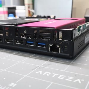 Peut inclure: Un châssis d'ordinateur noir doté d'une carte graphique rose proéminente installée sur le dessus. Le panneau arrière présente plusieurs ports d'entrée et de sortie, notamment des connexions USB, HDMI et Ethernet, alignés horizontalement.