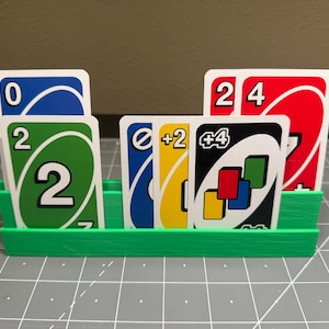 Puede incluir: Un soporte verde para cartas impreso en 3D lleno de cartas de Uno. Las cartas son de varios colores, incluyendo azul, verde, amarillo y rojo, con números e instrucciones especiales. El soporte está diseñado para mantener las cartas organizadas durante el juego.