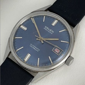 May include: Vintage Gruen Precision watch with a navy blue dial, silver hands, and a date window at the 3 o'clock position. The watch has a stainless steel case and a black leather strap. The dial reads "Gruen Precision", "Automatic", and "Incabloc".