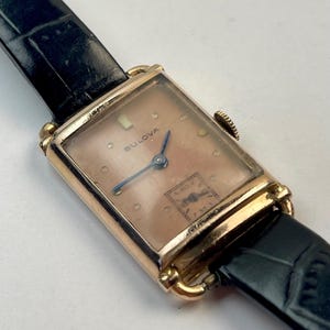 May include: Vintage Bulova watch with a rectangular gold-tone case and a pinkish-beige dial. The watch features blue hands and a black leather strap. The Bulova logo is visible on the watch face.