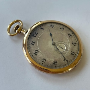 Puede incluir: Un reloj de bolsillo dorado con esfera crema y números negros. El reloj tiene un pequeño dial de segundos y una tija de cuerda. El reloj está sobre un fondo blanco.