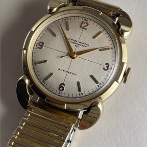 May include: A gold-toned wristwatch with a white face and black hands. The watch face has black Roman numeral hour markers and black markings for the minutes. The watch face also has the text "ULYSSE NARDIN CHRONOMETER CO. AUTOMATIC". The watch is on a gold-toned metal band.
