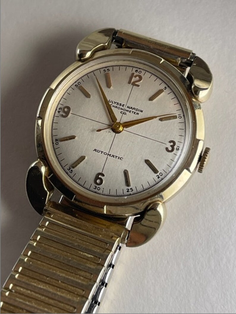 May include: A gold-toned wristwatch with a white face and black hands. The watch face has black Roman numeral hour markers and black markings for the minutes. The watch face also has the text "ULYSSE NARDIN CHRONOMETER CO. AUTOMATIC". The watch is on a gold-toned metal band.