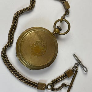 May include: A vintage brass pocket watch with an engraved face and chain. The watch has a detailed design on the cover and is connected to a chain with a T-bar clasp. The watch is a classic timepiece, a traditional accessory.