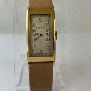 May include: A vintage Bulova wristwatch with a rectangular gold-tone case and a light-colored face. The watch features gold hands and numerals, with a brown leather band. The watch face displays the brand name and the word "SWISS".