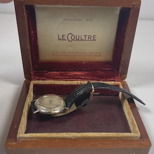 May include: A vintage Le Coultre watch with a silver-tone face and black leather band, displayed in a wooden box with a red velvet interior. The box lid features the text "Established 1833" and "LE COULTRE".