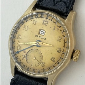 Może przedstawiać: Złoty zegarek na rękę z czarnym skórzanym paskiem. Tarcza zegarka ma złote tło z czarnymi rzymskimi cyframi godzinowymi i mniejszym tarczą na godzinie 3. Tarcza zegarka zawiera również tekst "BENRUS" i "SA".