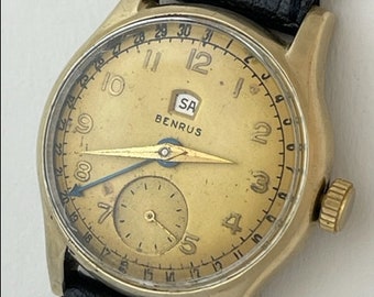 Puede incluir: Un reloj de pulsera dorado con una correa de cuero negro. La esfera del reloj tiene un fondo dorado con marcadores de hora en números romanos negros y un dial más pequeño en la posición de las 3 en punto. La esfera del reloj también presenta el texto "BENRUS" y "SA".