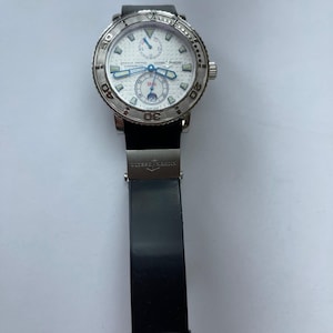 May include: A luxury wristwatch with a white face, silver bezel, and black band. The watch features blue hands and sub-dials, with the brand name "ULYSSE NARDIN" visible on the face. The band is black and made of rubber.