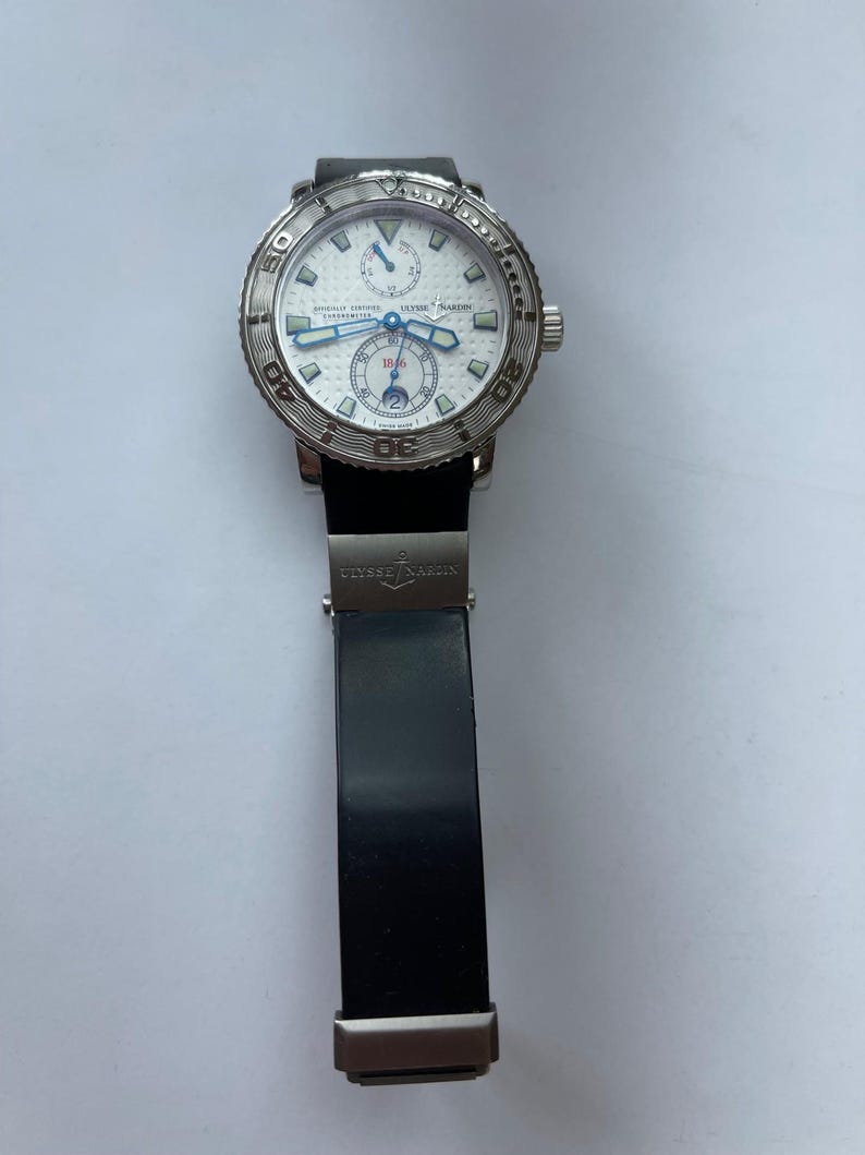 May include: A luxury wristwatch with a white face, silver bezel, and black band. The watch features blue hands and sub-dials, with the brand name "ULYSSE NARDIN" visible on the face. The band is black and made of rubber.