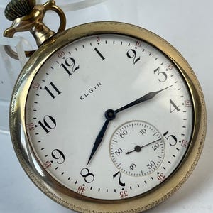 Puede incluir: Un reloj de bolsillo Elgin dorado con esfera blanca. El reloj presenta números negros, manecillas negras y un pequeño dial de segundos. La palabra "ELGIN" está impresa en la esfera. El reloj tiene una corona texturizada y un bisel detallado.
