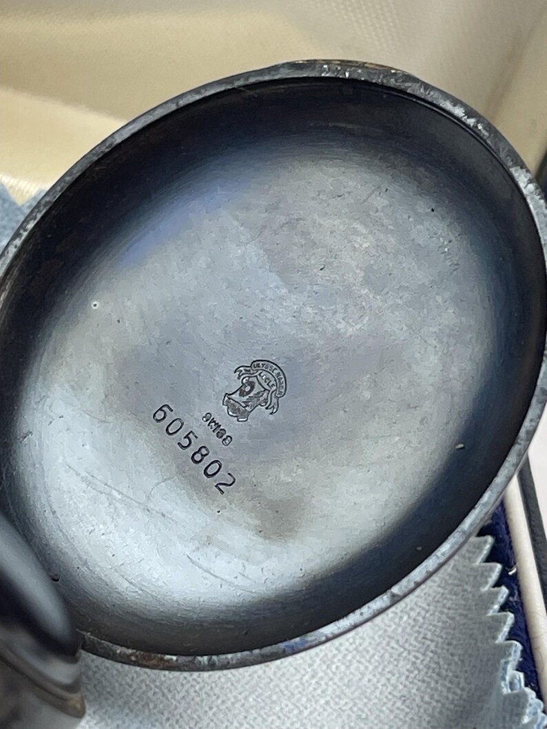 May include: The interior of a dark metallic pocket watch case featuring a circular emblem with the text Ulysse Nardin Le Locle, the word Swiss, and the engraved serial number 605802 centered on the smooth, slightly tarnished dark grey surface.