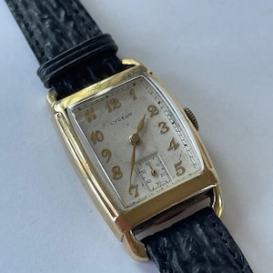 May include: A gold-toned wristwatch with a white face and black leather band. The watch face has black Roman numeral hour markers and a small seconds dial. The watch is labeled "LYCEUM" at the top.