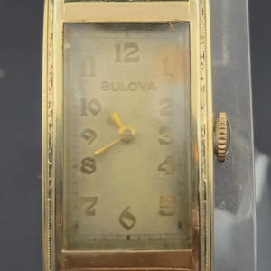 May include: A vintage Bulova wristwatch with a rectangular gold-toned case. The watch face is a light gold color with black numerals and hands. The watch has a small crown on the right side.