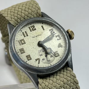 May include: A vintage Tavannes wristwatch with a weathered, off-white dial featuring black Arabic numerals and a small sub-dial for seconds. The watch has a dark, metallic case, a gold-toned crown, and a textured, olive-green woven fabric strap.