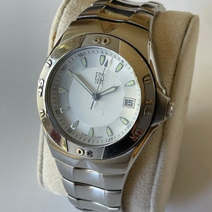 Peut inclure: Une montre-bracelet argentée avec un cadran blanc et des aiguilles noires. La montre a un bracelet en métal argenté et une fenêtre de date à 3 heures. Le cadran de la montre a des marques noires pour les heures et les minutes, et le texte "ESA Swiss Quartz" est imprimé sur le cadran.