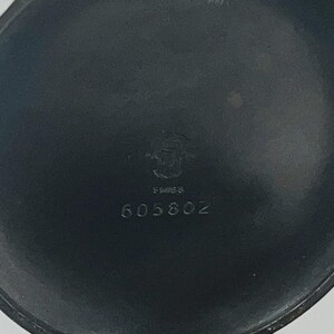 May include: A circular, matte black metal watch case back featuring a central engraved emblem, the word SWISS, and the numerical sequence 605802. The surface is smooth with a dark, uniform finish and a slightly raised outer rim edge.