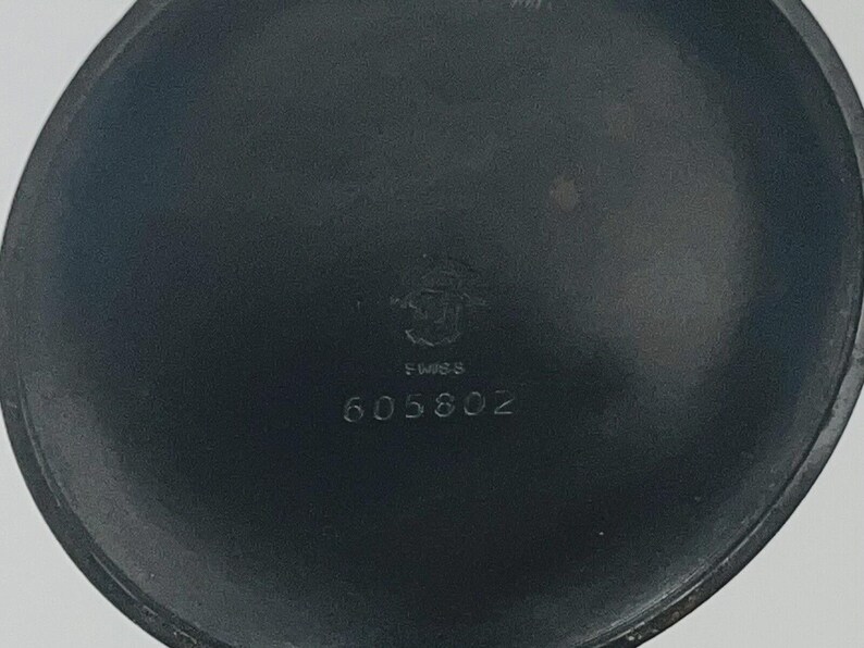 May include: A circular, matte black metal watch case back featuring a central engraved emblem, the word SWISS, and the numerical sequence 605802. The surface is smooth with a dark, uniform finish and a slightly raised outer rim edge.