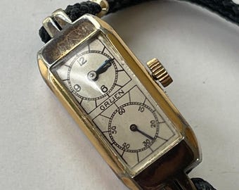 Può includere: Un orologio da polso Gruen dorato con un cinturino in corda nero. L'orologio ha due quadranti, uno con numeri romani e l'altro con numeri.