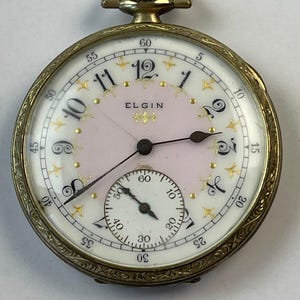 May include: Antique Elgin pocket watch with a gold-tone case and a white and pink face. The watch features black hands, gold-tone accents, and a subdial. The face displays the brand name "ELGIN". The watch has a detailed, engraved bezel.