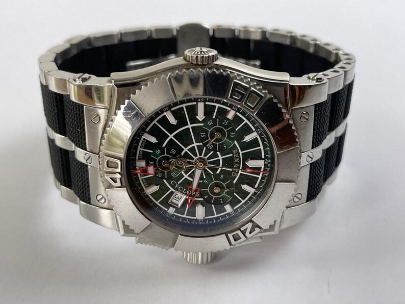 May include: A silver and black wristwatch with a green face and a black rubber strap. The watch has a date window and a chronograph function. The face of the watch has the word "Cyclops" printed on it.