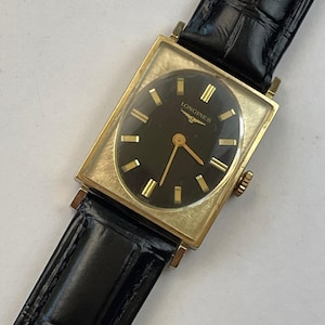 Könnte beinhalten: Eine Vintage-Longines-Uhr mit einem rechteckigen, goldfarbenen Gehäuse und einem schwarzen Zifferblatt. Das Zifferblatt hat goldfarbene Stundenmarkierungen und Zeiger. Die Uhr hat ein schwarzes Lederarmband.
