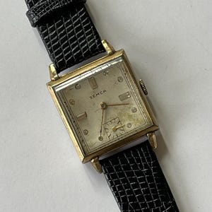 May include: A gold-toned square wristwatch with a white face and black leather band. The watch face has a silver-toned hour and minute hand, a black second hand, and the word "SEMCA" printed in black.