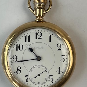 Puede incluir: Un reloj de bolsillo dorado con esfera blanca y números negros. La esfera del reloj muestra la marca "HOWARD" y una esfera secundaria más pequeña. El reloj tiene una corona y un lazo para una cadena.