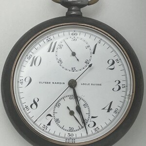 May include: A Ulysse Nardin Locle Suisse pocket watch with a white dial, black Arabic numerals, and two sub-dials. The watch features a dark metal case, a silver-toned crown with a circular loop, and black hands indicating the time.