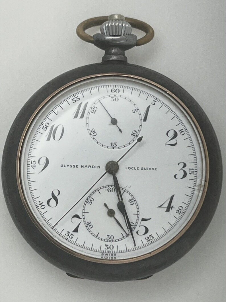 May include: A Ulysse Nardin Locle Suisse pocket watch with a white dial, black Arabic numerals, and two sub-dials. The watch features a dark metal case, a silver-toned crown with a circular loop, and black hands indicating the time.