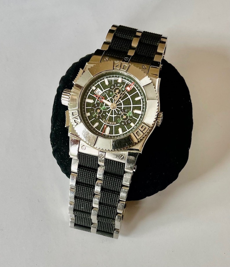 May include: A silver and black wristwatch with a green face and a black rubber strap. The watch has a chronograph function and a date window. The watch face has the word "CHRONO" printed on it.