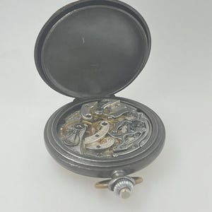May include: A dark gray metal pocket watch with an open case back, revealing the intricate mechanical movement inside. The watch features a circular frame, a winding crown at the top, and a hinged lid that is positioned upright behind the mechanism.
