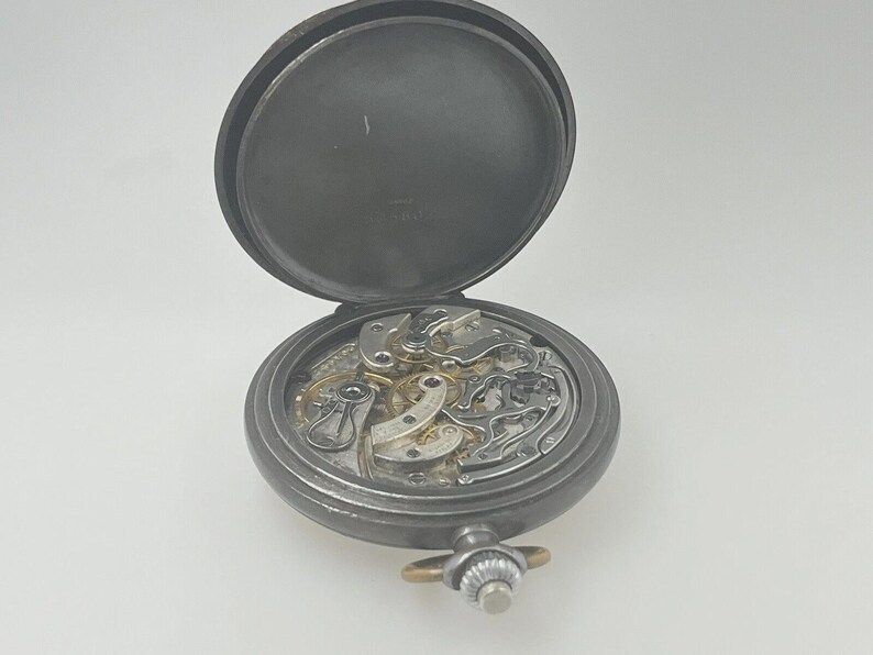 May include: A dark gray metal pocket watch with an open case back, revealing the intricate mechanical movement inside. The watch features a circular frame, a winding crown at the top, and a hinged lid that is positioned upright behind the mechanism.